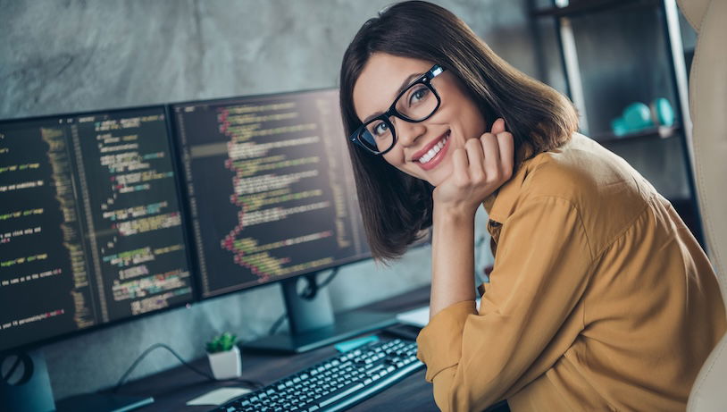 Woman with glasses smiling at camera with a keyboard and two screens with a code editor open