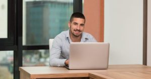 Happy Male Student In Library With Laptop A male behind a laptop at a desk in front of a window