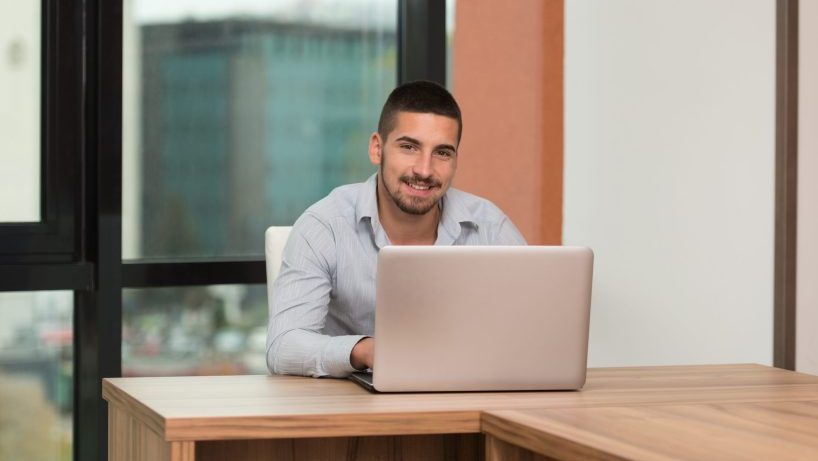 A male behind a laptop at a desk in front of a window